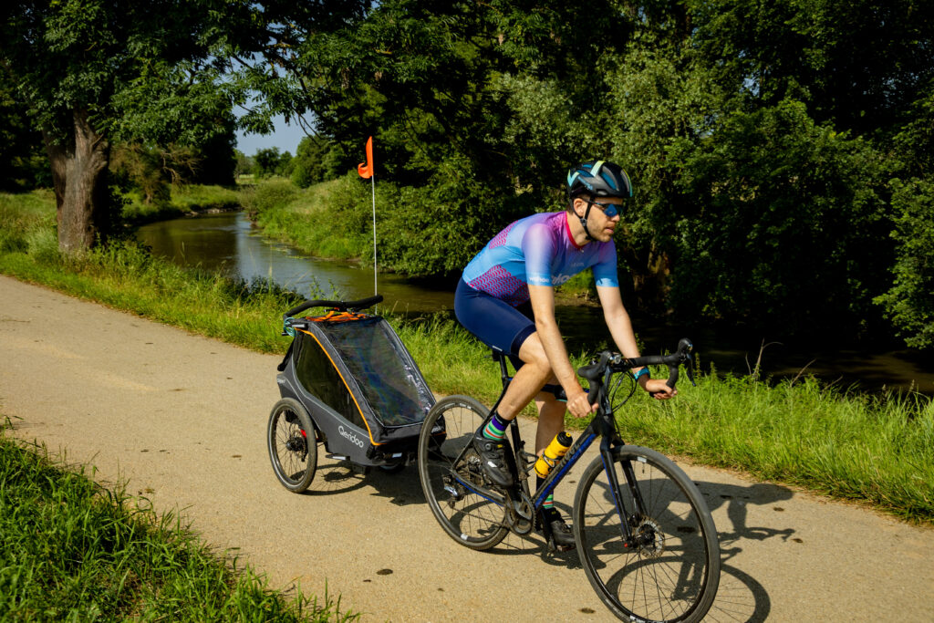 Konstantin (aka The Cycling Dad) on riverside trail with Qeridoo Kidgoo 1 trailer trailer near trees and greenery